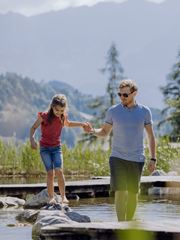 A girl balancing on rocks at the natural swimming pond, with an adult holding her hand, surrounded by green nature in summer.