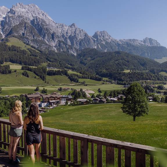 Zwei Frauen beim Wandern im Sommer, blicken auf das Hotel Puradies, umgeben von grünen Wiesen und der Bergkulisse im Hintergrund.