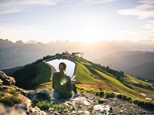 Person sitzt beim Wandern auf Felsen mit Bergpanorama und Sonnenaufgang in Leogang Person sitzt beim Wandern auf Felsen mit Bergpanorama und Sonnenaufgang in Leogang