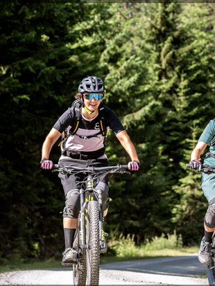 Zwei lachende Mountainbikerinnen fahren auf einem Forstweg im Wald bei Leogang