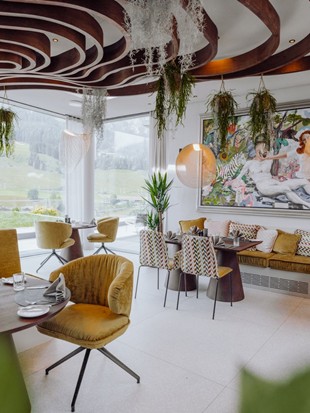 Schön gedeckte Tische im Gourmetrestaurant Essenz, umgeben von stilvollem Ambiente und einer Glasfront mit Blick auf die sommerliche Landschaft und die Bergkulisse.