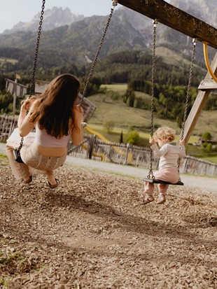 Eine Frau und ein kleines Kind hutschen im Sommer auf dem Abenteuerspielplatz des Puradies, mit grünen Wiesen und Bergen im Hintergrund.