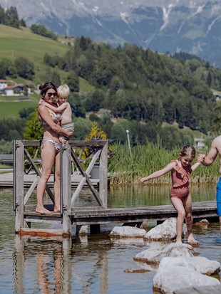 Eine Frau hebt einen kleinen Jungen und steht am Steg des Naturbadeteichs, während ein Mann einem kleinen Mädchen beim Balancieren über die Steine hilft, im Sommer umgeben von grüner Landschaft.