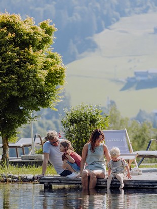 Zwei Erwachsene und zwei Kinder sitzen im Sommer am Steg des Naturbadeteichs und baden ihre Füße, mit grünen Wiesen im Hintergrund.