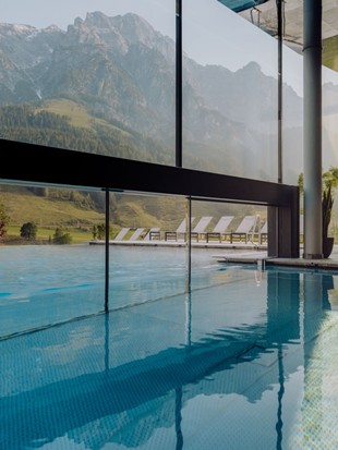 heaven-spa-puradies-credit-thecreatingclick Infinity pool from inside, with a view through the glass front of the summer landscape and the mountain backdrop.