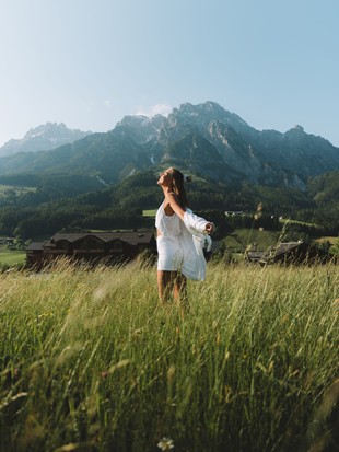 Eine Frau steht inmitten einer grünen Wiese im Sommer und genießt die Sonne, mit der Bergkulisse im Hintergrund.