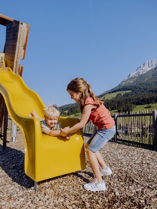 Kleines Mädchen hilft einem kleinen Jungen an der Rutsche des Abenteuerspielplatzes im Sommer, umgeben von grünen Wiesen und Bergen im Hintergrund.