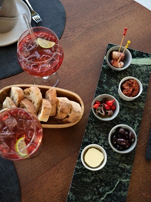 Tapas Experience im Restaurant Ess:enz – Puradies Leogang Tapas-Auswahl mit Oliven, Peperoni und Dips auf grünem Marmor, frisches Weißbrot und zwei Spritz-Cocktails mit Eiswürfeln und Limette im Restaurant Ess:enz, Naturresort Puradies Leogang
