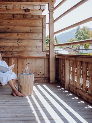 Eine Frau sitzt in einem Holzstuhl auf dem Balkon des Zimmers und liest eine Zeitung.