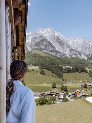 Eine Frau von hinten schaut von ihrem Zimmerbalkon auf die grüne, sommerliche Landschaft und die Bergkulisse.