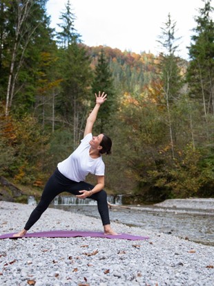 Sabine Breitsameter, die das Yoga-, Wein- und Genuss-Retreat durchführt, macht Yoga auf einer Yogamatte auf einem Schotterplatz neben einem Fluss im Wald.