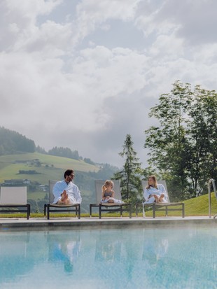Zwei Erwachsene und ein kleines Mädchen liegen auf den Liegen neben dem Infinity-Pool des Heaven Spas im Sommer, mit grüner Landschaft im Hintergrund.