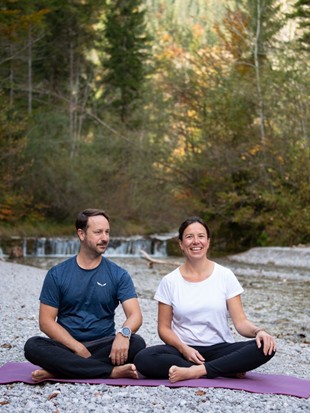Ein Mann und eine Frau, Matthias und Sabine Breitsameter, die das Yoga-, Wein- und Genuss-Retreat durchführen, sitzen auf Yogamatten auf einem Schotterplatz neben einem Fluss inmitten eines Waldes