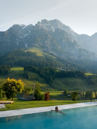 A person in the outdoor infinity pool, surrounded by green meadows and mountains in the background.