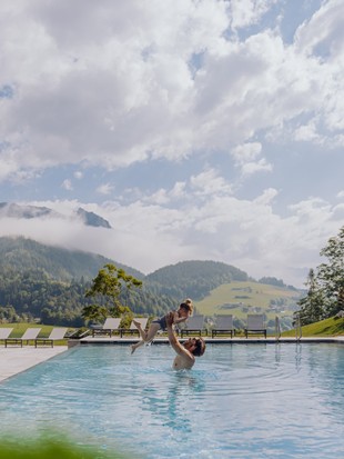 Ein Erwachsener hebt ein Kind im Infinity Pool des Naturresorts Puradies im Sommer hoch, umgeben von grüner Landschaft.