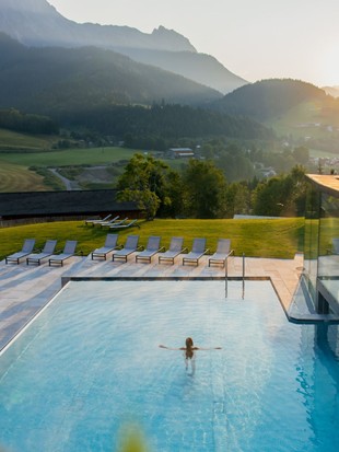 A person in the outdoor infinity pool, surrounded by green meadows and mountains in the background, with a summer atmosphere.