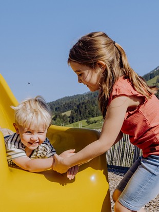Ein kleines Mädchen hilft einem kleinen Jungen beim Runterrutschen auf der Rutsche am Abenteuerspielplatz im Sommer, mit grünen Wiesen im Hintergrund.