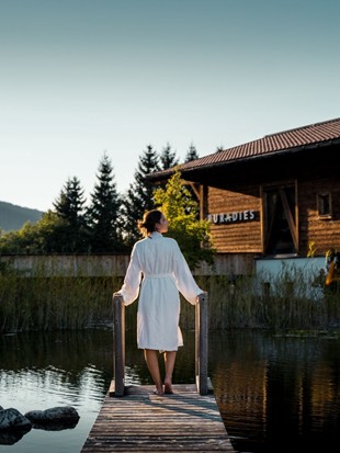 Eine Frau steht im Bademantel am Steg des Naturbadeteichs im Sommer bei Sonnenuntergang.