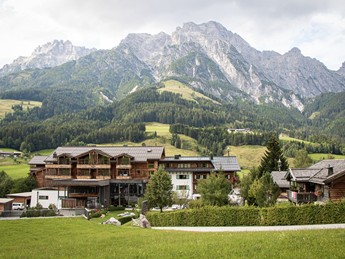 Front view of Hotel Puradies in summer with a green meadow in the foreground and mountains in the background.
