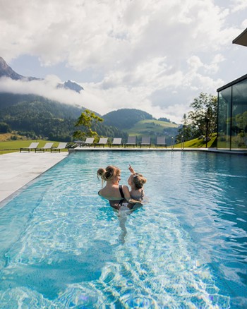 Frau steht im Infinity-Pool im Freien, mit einem kleinen Kind auf dem Arm, im Sommer, mit grünen Bergen im Hintergrund.