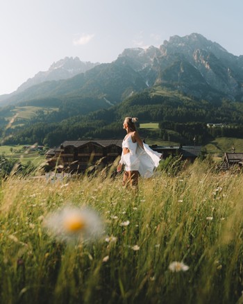 Frau steht im Sommer inmitten einer grünen Wiese, mit dem Hotel Puradies und den Bergen im Hintergrund.