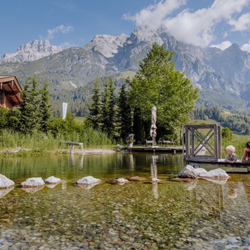 Eine Frau und ein Kind liegen am Steg des Naturbadeteichs und schauen in den See. Im Hintergrund sind das Badhaus, grüne Wiesen und die Berglandschaft zu sehen, alles in einer sommerlichen Atmosphäre.