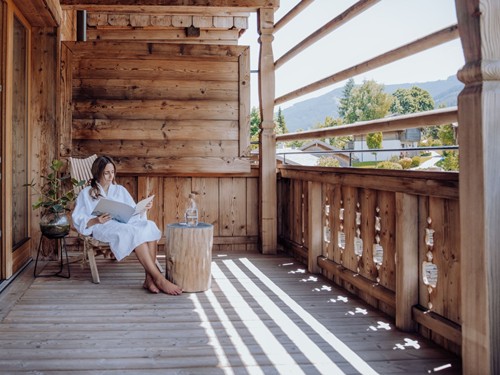 Frau im Bademantel sitzt lesend auf dem Holzbalkon mit Bergblick im Naturresort Puradies in Leogang.
