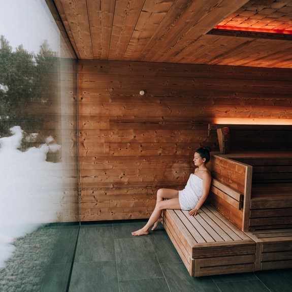 Frau sitzt in der Holzsauna und schaut durch die große Glasfront nach draußen auf die Schneelandschaft.