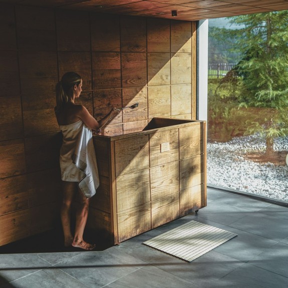 Frau steht in der Holzsauna und schaut durch die Glasfront auf die sommerliche Landschaft.