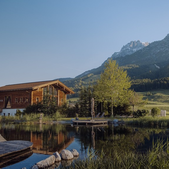 Naturbadeteich und Badhaus im Naturresort Puradies, mit Bergen im Hintergrund und grünen Wiesen im Sommer.