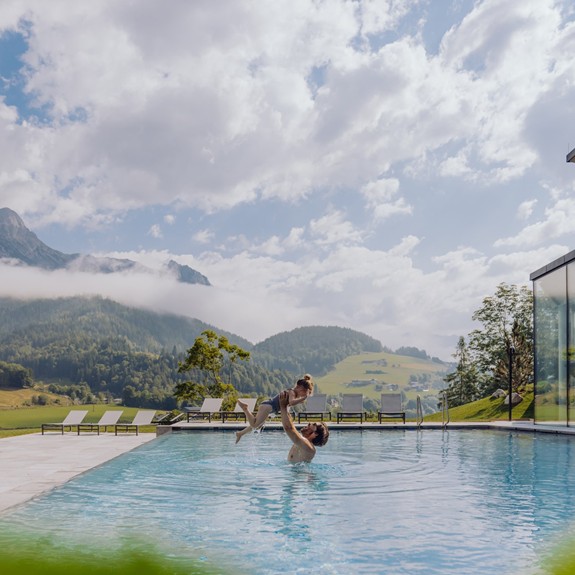 Ein Erwachsener hebt ein Kind im Infinity Pool des Naturresorts Puradies im Sommer hoch, umgeben von grüner Landschaft.