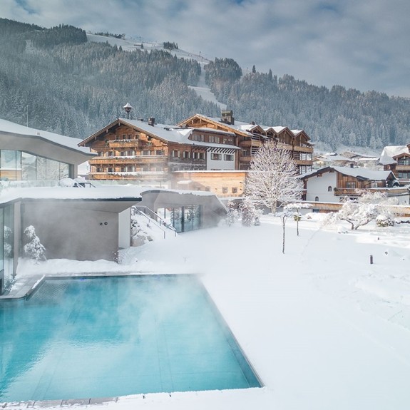 Luftaufnahme des Infinity-Pools im Winter, umgeben von Schnee, mit Hotelanlage im Hintergrund.