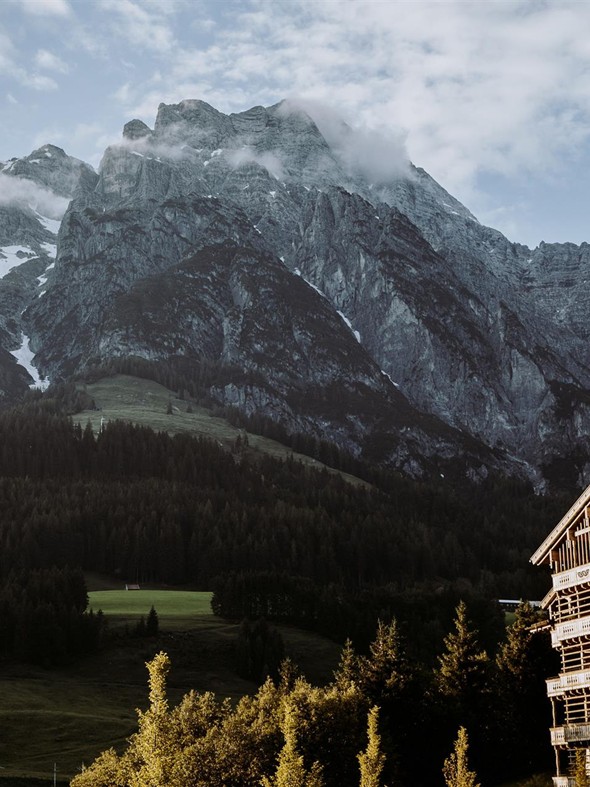 Part of the hotel with a natural wooden facade in summer, surrounded by green trees, meadows, and mountains in the background.