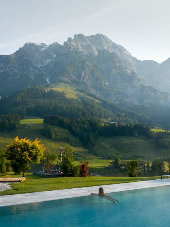 Person im Infinity-Pool im Freien, umgeben von grünen Wiesen und Bergen im Hintergrund.